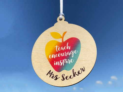 Wooden keepsake with white satin ribbon, printed apple shape with rainbow colour. Inside the wording 'teach, encourage, inspire'.